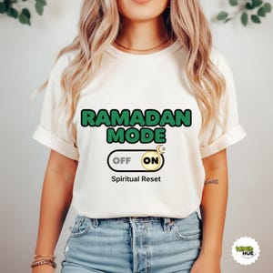 May include: Cream-colored t-shirt with the words "RAMADAN MODE" in green and black. Below the text is a switch graphic with "OFF" and "ON" options, and the words "Spiritual Reset". The shirt has rolled-up sleeves.