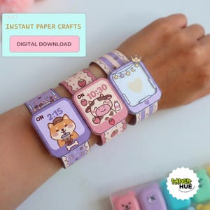 Puede incluir: Tres relojes de pulsera de papel en tonos morados y rosas con ilustraciones de dibujos animados de un perro con una bebida boba, una vaca con fresas y un icono de corazón. Las esferas muestran las horas 2:15 y 10:30.
