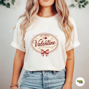 May include: Cream-colored t-shirt with a Valentine's Day design. The design features the words "My Sweet Valentine" in a script font, along with a bow and hearts. The shirt is made of a soft material and has rolled-up sleeves.