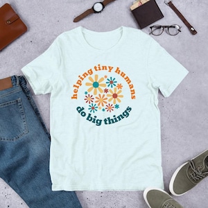 May include: A light blue t-shirt with a graphic design of colourful flowers and the text "helping tiny humans do big things".