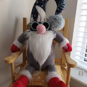 May include: A whimsical gnome doll with a long, curved hat featuring a polar bear and snowflake pattern. The gnome wears blue sunglasses and has a pink nose, a long white beard, and red boots. It is seated on a yellow floral cushion on a wooden chair.