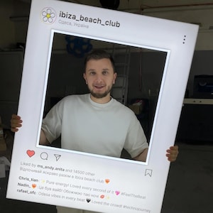 May include: A large, white Instagram-style photo frame with the text "ibiza_beach_club" and "Odesa, Україна" at the top. The frame has a heart, comment, and send icons. A person is holding the frame, smiling. Text includes comments and hashtags.