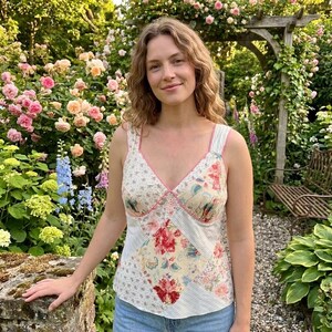 Pode incluir: Uma blusa sem mangas com um design de patchwork de vários padrões florais em tons de vermelho, rosa, amarelo e azul sobre um fundo creme. A peça tem um decote em V com um rebordo fino cor-de-rosa e alças largas nos ombros.