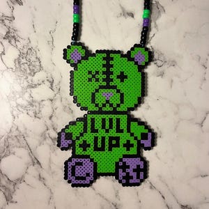 May include: A green and black beaded necklace featuring a bear pendant. The bear has purple accents on its ears, paws, and nose, and the text "LIL UP+" is displayed on its chest. The necklace has a black, green, and purple beaded chain.