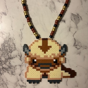 May include: A necklace featuring a pixelated pendant made of fused plastic beads shaped like a creature with cream-colored fur, a brown arrow on its head, and dark grey horns. The necklace cord consists of alternating gold, black, and brown beads.