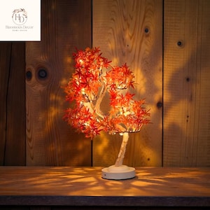 Puede incluir: Lámpara decorativa LED con forma de árbol, hojas naranjas vibrantes y base blanca. Las ramas están iluminadas con pequeñas luces, creando un brillo cálido. La lámpara se encuentra sobre un fondo de madera, añadiendo un toque natural a la decoración.