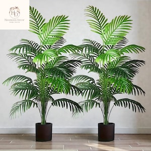 Könnte beinhalten: Zwei künstliche Palmen mit üppigen grünen Wedeln und dunkelbraunen Töpfen. Die Pflanzen haben mehrere Stämme und ein volles, realistisches Aussehen, das jedem Innenraum einen Hauch von Natur verleiht. Das Logo "Havenous Decor HOME DECOR" befindet sich oben links.