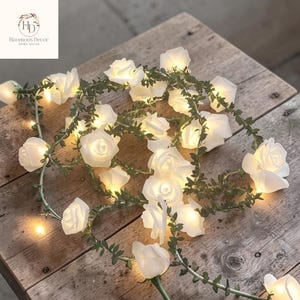 May include: A string of white rose-shaped fairy lights with green leaves, arranged on a rustic wooden surface. The lights are illuminated, creating a soft glow. The product is from Havenous Decor.