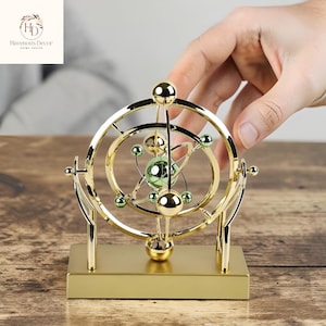 Könnte beinhalten: Eine weiße Keramiktasse mit schwarzem Henkel und dem Text "But First, Coffee" in fetter, schwarzer Schrift. Die Tasse steht vor einem verschwommenen Hintergrund, der eine gemütliche Umgebung suggeriert.