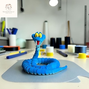 May include: A blue snake figurine with yellow eyes and a red mouth. The snake is coiled and has a textured surface. The figurine is on a gray surface. The logo for Havenous Decor is in the background.