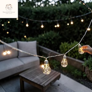 Peut inclure: Une guirlande d'ampoules en verre transparent avec des culots argentés, contenant chacune de petites lumières féeriques. Les ampoules sont reliées par un fin fil blanc et sont allumées, diffusant une lumière chaude dans l'obscurité.