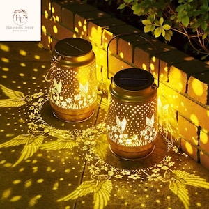 以下が含まれることがあります： ハチドリと花のカットアウトデザインが施された、2つの金色のソーラーランタン。ランタンにはハンドルとソーラーパネルが付いています。ランタンからの光は地面に装飾的なパターンを投影し、暖かく居心地の良い雰囲気を作り出しています。