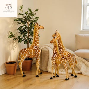 May include: Three plush giraffe figurines with brown and white patterned hides and long necks. The giraffes vary in size, with the tallest standing near a potted plant. The figurines are placed on a light wood floor.