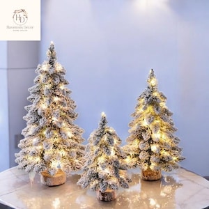 May include: Three artificial Christmas trees with a snowy effect and warm white lights. The trees are set on wooden bases and are arranged on a marble-like surface. The trees vary in size.