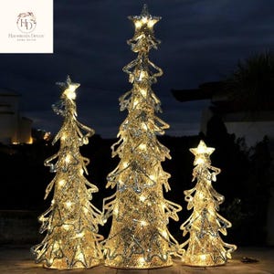 Puede incluir: Tres árboles de Navidad decorativos iluminados de diferentes tamaños. Cada árbol dorado tiene una estrella en la parte superior y está iluminado con luces blancas cálidas. Los árboles están sobre un fondo oscuro.
