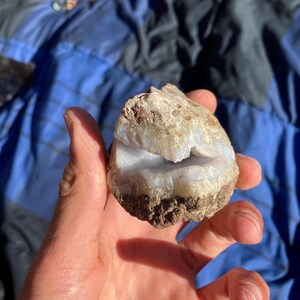 May include: A hand holding a geode with a light blue interior and a brown and beige exterior. The geode has a rough, natural texture and a visible cavity. The background is a blue and black patterned fabric.