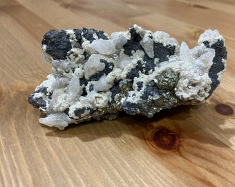 May include: A cluster of white crystals embedded in a dark gray rock with a metallic sheen. The rock has a rough, uneven surface.
