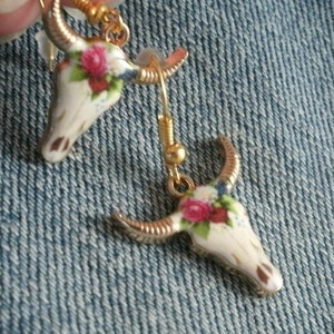 May include: A pair of gold tone earrings with a white porcelain skull design. The skull is decorated with pink and white flowers.