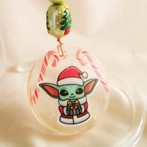 Peut inclure: Une d&eacute;coration de No&euml;l repr&eacute;sentant un personnage vert et blanc portant un bonnet de P&egrave;re No&euml;l et tenant un cadeau. La d&eacute;coration est orn&eacute;e de cannes en sucre et d'un n&oelig;ud en tartan rouge et vert.