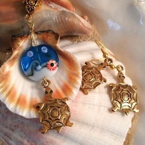 May include: A gold necklace with a blue and white glass turtle pendant, and matching gold turtle earrings. The jewelry is displayed on a seashell background.