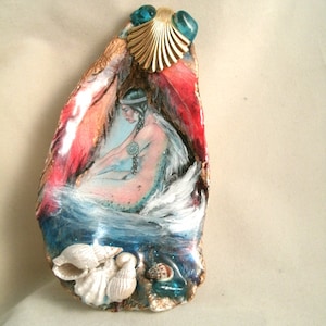 May include: A painted oyster shell with a mermaid figure inside, surrounded by seashells and blue glitter. The shell is decorated with a gold seashell and blue glass beads.