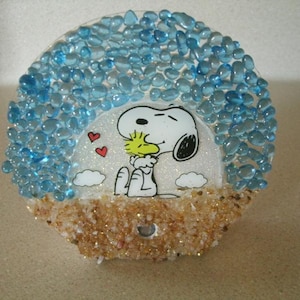 May include: A Snoopy-themed decorative piece featuring a cartoon Snoopy hugging Woodstock, surrounded by blue and tan decorative stones. The artwork includes red hearts and white clouds, creating a charming, whimsical design.