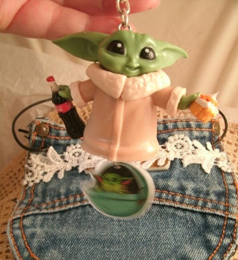 Op de afbeelding: Een groen en bruin beeldje van een personage uit de Star Wars-franchise, dat een fles Coca-Cola en een bakje met eten vasthoudt. Het beeldje is bevestigd aan een sleutelhanger.