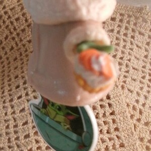 Op de afbeelding: Een keramiek beeldje van een groen buitenaards wezen dat een witte gewatteerde jas draagt en een klein oranje voorwerp vasthoudt. Het beeldje staat op een gehaakt onderzetter.