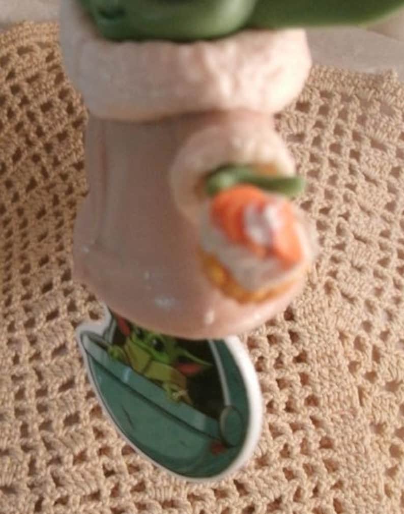 Op de afbeelding: Een keramiek beeldje van een groen buitenaards wezen dat een witte gewatteerde jas draagt en een klein oranje voorwerp vasthoudt. Het beeldje staat op een gehaakt onderzetter.