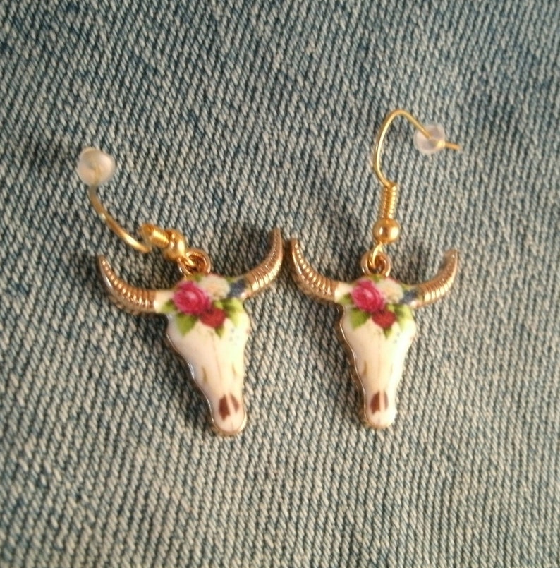 May include: A pair of gold tone earrings with a white skull design and floral accents. The skull is decorated with pink, red, and green flowers.