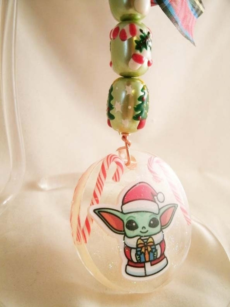Peut inclure: Une boule de No&euml;l ronde transparente avec un motif de canne en sucre verte et blanche. La boule pr&eacute;sente une image de dessin anim&eacute; d'un extraterrestre vert portant un chapeau de P&egrave;re No&euml;l et tenant un cadeau.