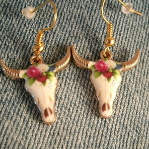 May include: A pair of gold tone earrings with a white porcelain bull skull design. The skull is decorated with a floral pattern in shades of pink, red, and blue.