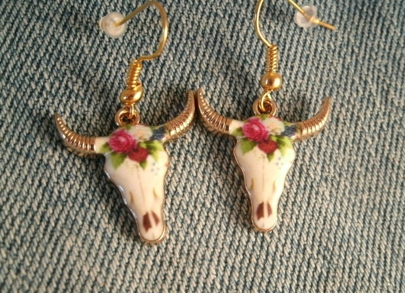 May include: A pair of gold tone earrings with a white porcelain bull skull design. The skull is decorated with a floral pattern in shades of pink, red, and blue.