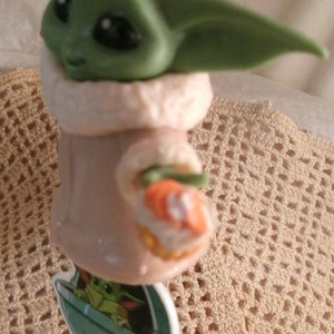 Op de afbeelding: Een groen en bruin beeldje van een personage uit de Star Wars-franchise, bekend als "The Child" of "Baby Yoda". Het beeldje draagt een bruine gewatteerde mantel en houdt een klein oranje voorwerp in zijn hand. Het beeldje is bevestigd aan een sleutelhanger.