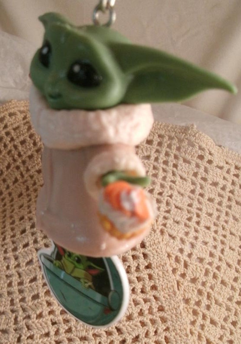 Op de afbeelding: Een groen en bruin beeldje van een personage uit de Star Wars-franchise, bekend als "The Child" of "Baby Yoda". Het beeldje draagt een bruine gewatteerde mantel en houdt een klein oranje voorwerp in zijn hand. Het beeldje is bevestigd aan een sleutelhanger.