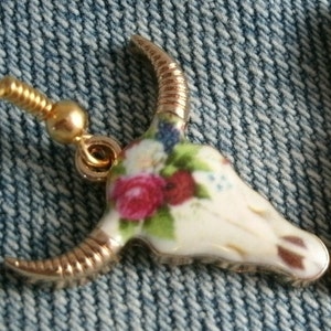 May include: Gold tone earrings with a white porcelain bull skull design. The skull is decorated with a floral pattern in shades of pink, red, and green.