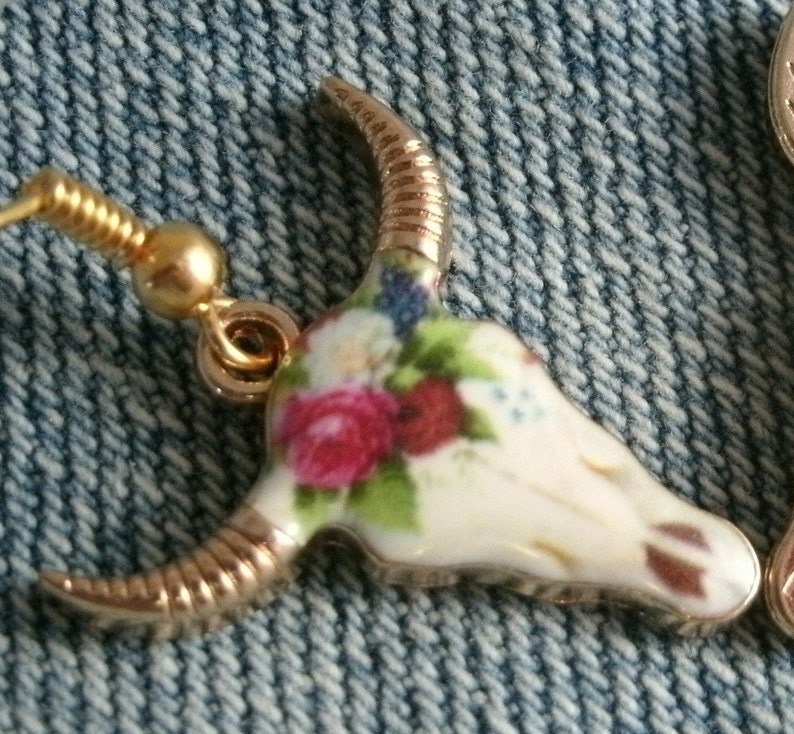 May include: Gold tone earrings with a white porcelain bull skull design. The skull is decorated with a floral pattern in shades of pink, red, and green.