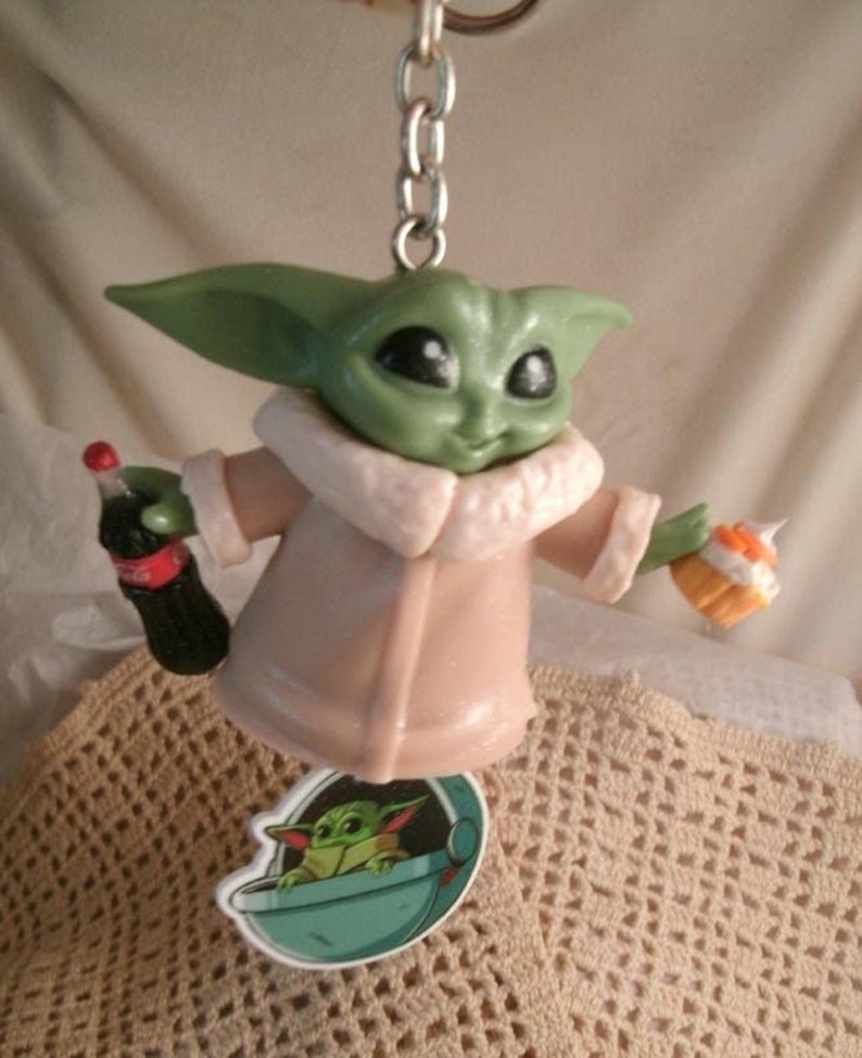 Op de afbeelding: Een groene en bruine plastic sleutelhanger met een personage uit de Star Wars-franchise, The Mandalorian. Het personage houdt een klein flesje frisdrank in de ene hand en een klein taartje in de andere. De sleutelhanger bevat ook een kleine sticker van het personage in een ruimteschip.