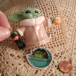 Op de afbeelding: Een groen en bruin beeldje van een personage uit de Star Wars-franchise, dat een klein flesje frisdrank en een cupcake vasthoudt. Het beeldje draagt een bruine jas met een roze bontkraag. Het beeldje is bevestigd aan een sleutelhanger met een kleiner beeld van het personage in een pod.
