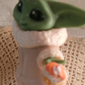 Op de afbeelding: Een groen en bruin beeldje van een personage uit de Star Wars-serie, met een witte en bruine jas. Het personage houdt een klein, oranje en wit voorwerp in zijn hand.