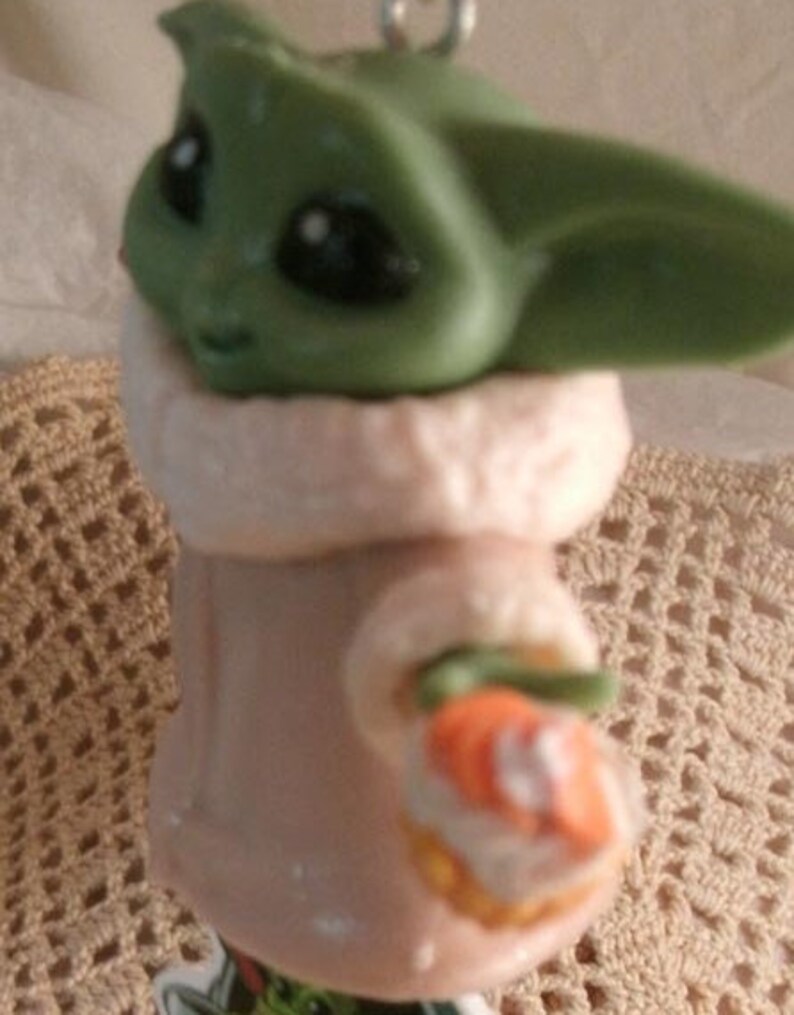 Op de afbeelding: Een groen en bruin beeldje van een personage uit de Star Wars-serie, met een witte en bruine jas. Het personage houdt een klein, oranje en wit voorwerp in zijn hand.