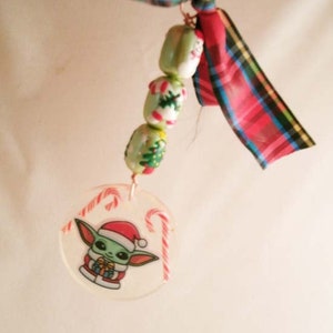 Peut inclure: Une d&eacute;coration de No&euml;l repr&eacute;sentant un personnage vert portant un bonnet de P&egrave;re No&euml;l rouge et tenant un cadeau, entour&eacute; de cannes en sucre. La d&eacute;coration est attach&eacute;e &agrave; une cha&icirc;ne de perles vertes et blanches avec des motifs de No&euml;l. Un ruban rouge et noir &agrave; carreaux est nou&eacute; au sommet de la d&eacute;coration.