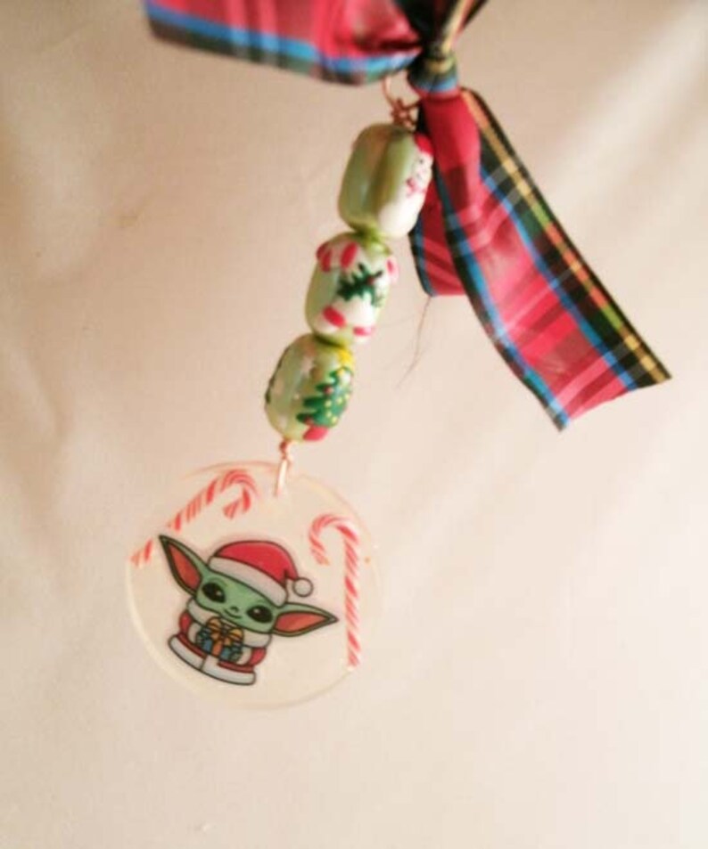 Peut inclure: Une d&eacute;coration de No&euml;l repr&eacute;sentant un personnage vert portant un bonnet de P&egrave;re No&euml;l rouge et tenant un cadeau, entour&eacute; de cannes en sucre. La d&eacute;coration est attach&eacute;e &agrave; une cha&icirc;ne de perles vertes et blanches avec des motifs de No&euml;l. Un ruban rouge et noir &agrave; carreaux est nou&eacute; au sommet de la d&eacute;coration.
