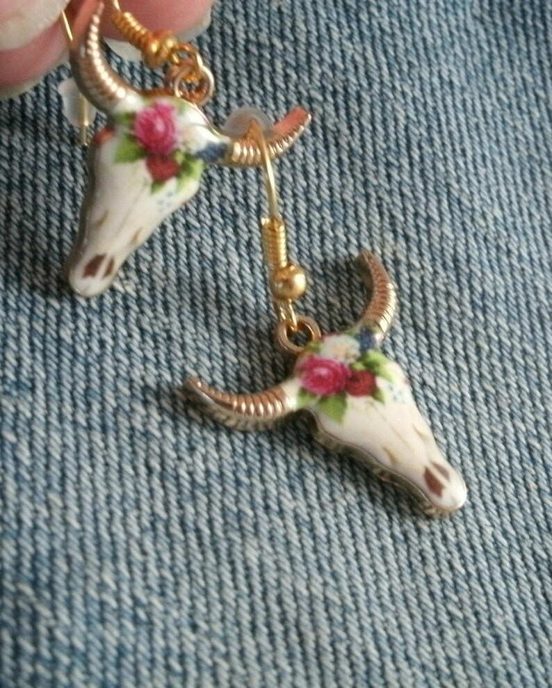 May include: A pair of gold tone earrings with a white porcelain skull design. The skull is decorated with pink and white flowers.