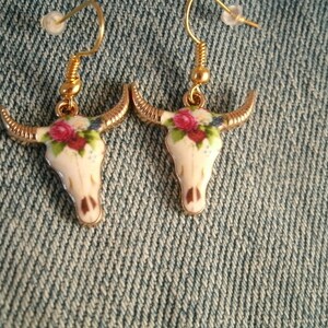 May include: A pair of gold tone earrings with a white porcelain bull skull design. The skull is decorated with pink and white flowers.