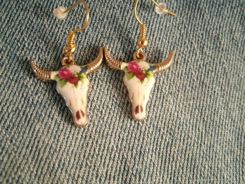 May include: A pair of gold tone earrings with a white porcelain bull skull design. The skull is decorated with pink and white flowers.