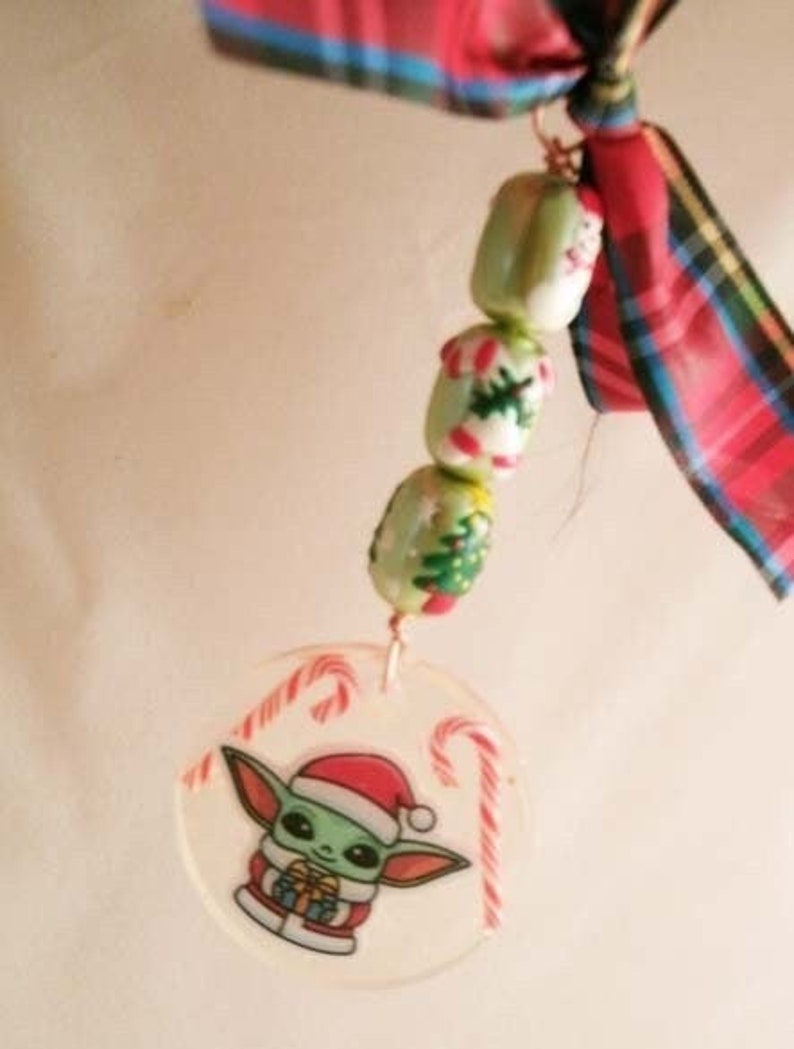 Peut inclure: Une d&eacute;coration de No&euml;l repr&eacute;sentant un personnage vert portant un bonnet de P&egrave;re No&euml;l et tenant un cadeau. La d&eacute;coration est entour&eacute;e de motifs de cannes en sucre et est attach&eacute;e &agrave; une cha&icirc;ne de perles aux motifs de No&euml;l.