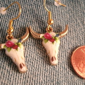 May include: A pair of gold tone earrings with white porcelain bull skull charms. The charms are decorated with pink and white roses and blue accents. The earrings are shown next to a US penny for size comparison.