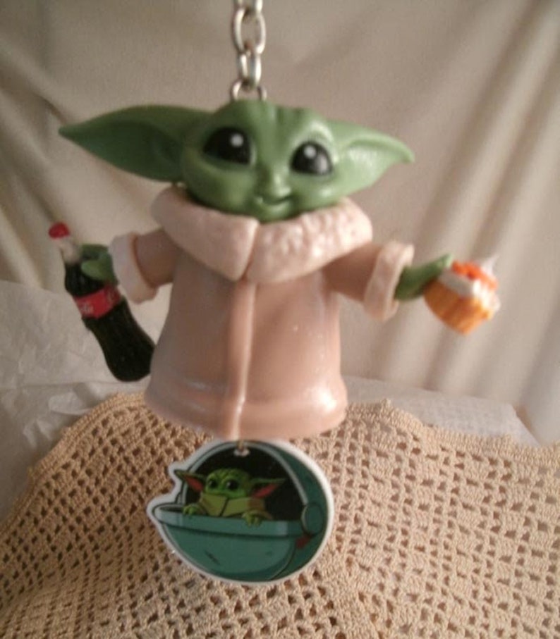 Op de afbeelding: Een groen en bruin beeldje van een personage uit de Star Wars-franchise, dat een fles frisdrank en een klein taartje vasthoudt. Het beeldje draagt een beige jas met een witte bontkraag. Het beeldje is bevestigd aan een sleutelhanger met een klein beeld van het personage in een ruimteschip.