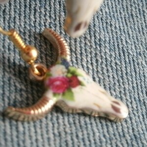 May include: Gold tone earrings with a white porcelain skull charm decorated with pink and blue floral details.